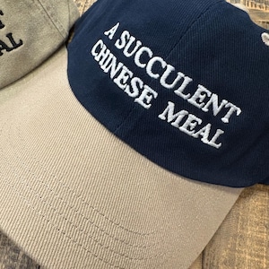 Puede incluir: Una gorra de b&eacute;isbol azul marino y beige con el texto "A Succulent Chinese Meal" bordado en blanco en la parte delantera.