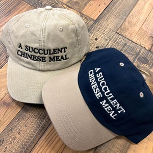 Puede incluir: Dos gorras de b&eacute;isbol con el texto "A succulent Chinese meal" bordado en la parte delantera. Una gorra es beige con una visera beige y la otra es azul marino con una visera beige.