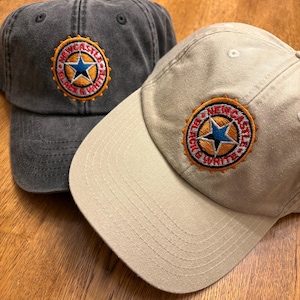 May include: Two baseball caps, one in charcoal grey and one in khaki, are shown. Each cap has an embroidered patch with a blue star and the words "Newcastle Black & White" in a circular design. The caps are on a wooden surface.