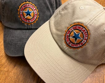 Puede incluir: Se muestran dos gorras de béisbol, una gris antracita y otra caqui. Cada gorra presenta un parche bordado con una estrella azul y las palabras "Newcastle Black & White" en un diseño circular. Las gorras están sobre una superficie de madera.