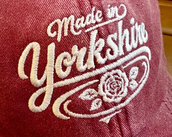 Puede incluir: Gorra de béisbol granate con texto bordado en blanco "Made in Yorkshire". El diseño incluye un emblema de rosa blanca. La gorra tiene una visera curva y un botón en la parte superior.