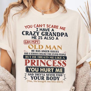 Op de afbeelding: Crèmekleurig T-shirt met een humoristische boodschap in rode, marineblauwe en zwarte tekst. De tekst luidt: "Je kunt me niet bang maken, ik heb een gekke opa..." De boodschap van het shirt is een speelse verklaring.