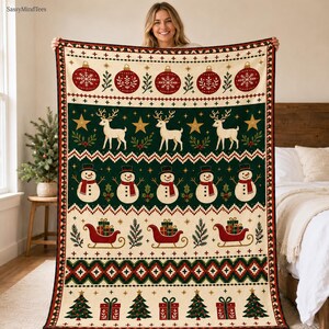 Könnte beinhalten: Eine rechteckige Wohndecke mit festlichem Weihnachtsmuster, bestehend aus horizontalen Bändern mit roten Christbaumkugeln, weißen Rentieren, Schneemännern mit roten Schals, roten Schlitten mit Geschenken und grünen Tannen auf cremefarbenem Grund.