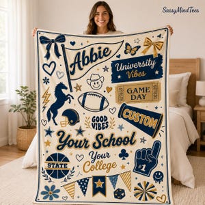 Peut inclure: Un plaid couleur crème avec des motifs bleu marine et dorés représentant un cheval, un ballon de football, un mégaphone, un doigt en mousse, des nœuds et des papillons. Le texte inclut "Abbie", "University Vibes", "GAME DAY", "GOOD VIBES", "Your School" et "Your College".