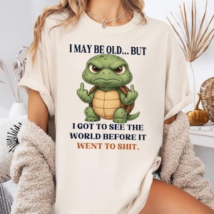 May include: Cream-coloured t-shirt with the text "I MAY BE OLD... BUT I GOT TO SEE THE WORLD BEFORE IT WENT TO SHIT." The design includes a cartoon turtle with an angry face, displaying the middle finger.