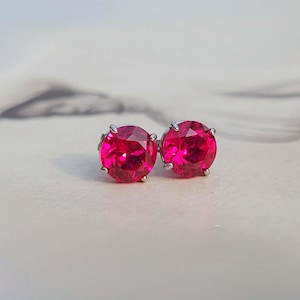May include: A pair of round, faceted, vibrant hot pink stud earrings set in silver-toned metal prongs. Each earring features a circular gemstone with a brilliant cut that reflects light, creating a clear, saturated color profile and a polished finish.
