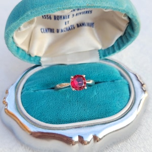 May include: A red gemstone ring in a teal velvet ring box. The box is labeled "1556 Royale 3-Rivieres et Centre d'Achats Banlieue".