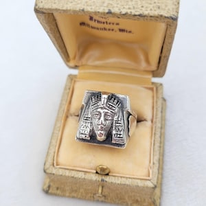 May include: A silver ring with a detailed pharaoh design, set in a vintage beige jewelry box. The ring features a sculpted face with intricate details and a red accent. The box is labeled with text.