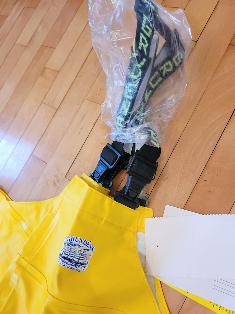 May include: Yellow Grund&eacute;ns foul-weather bib overalls with black suspenders. The overalls have a Grund&eacute;ns logo patch and are partially packaged in plastic. The suspenders are black with green text.