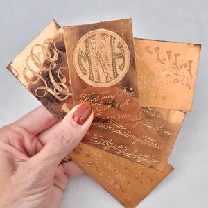Könnte beinhalten: Eine Sammlung kupferfarbener Metallkarten mit gravierten Texten und Designs. Eine Karte zeigt ein Monogramm in einem Kreis, andere handschriftliche Schriftzüge. Die Karten haben eine Vintage-Ästhetik.