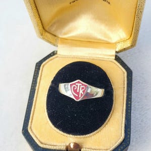 Puede incluir: Un anillo plateado con un emblema CTR rojo, presentado en un joyero vintage. La caja es dorada y negra, con el texto "E. A. Meckelberg Co. Jewelers Two Rivers, Wis." en la tapa interior.