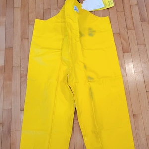 May include: Bright yellow waterproof bib overalls. The overalls have a high bib, suspenders, and straight legs. The garment is made of a shiny, waterproof material. The top of the overalls has a black strap and a clear plastic bag with the word "DENZ" printed on it.
