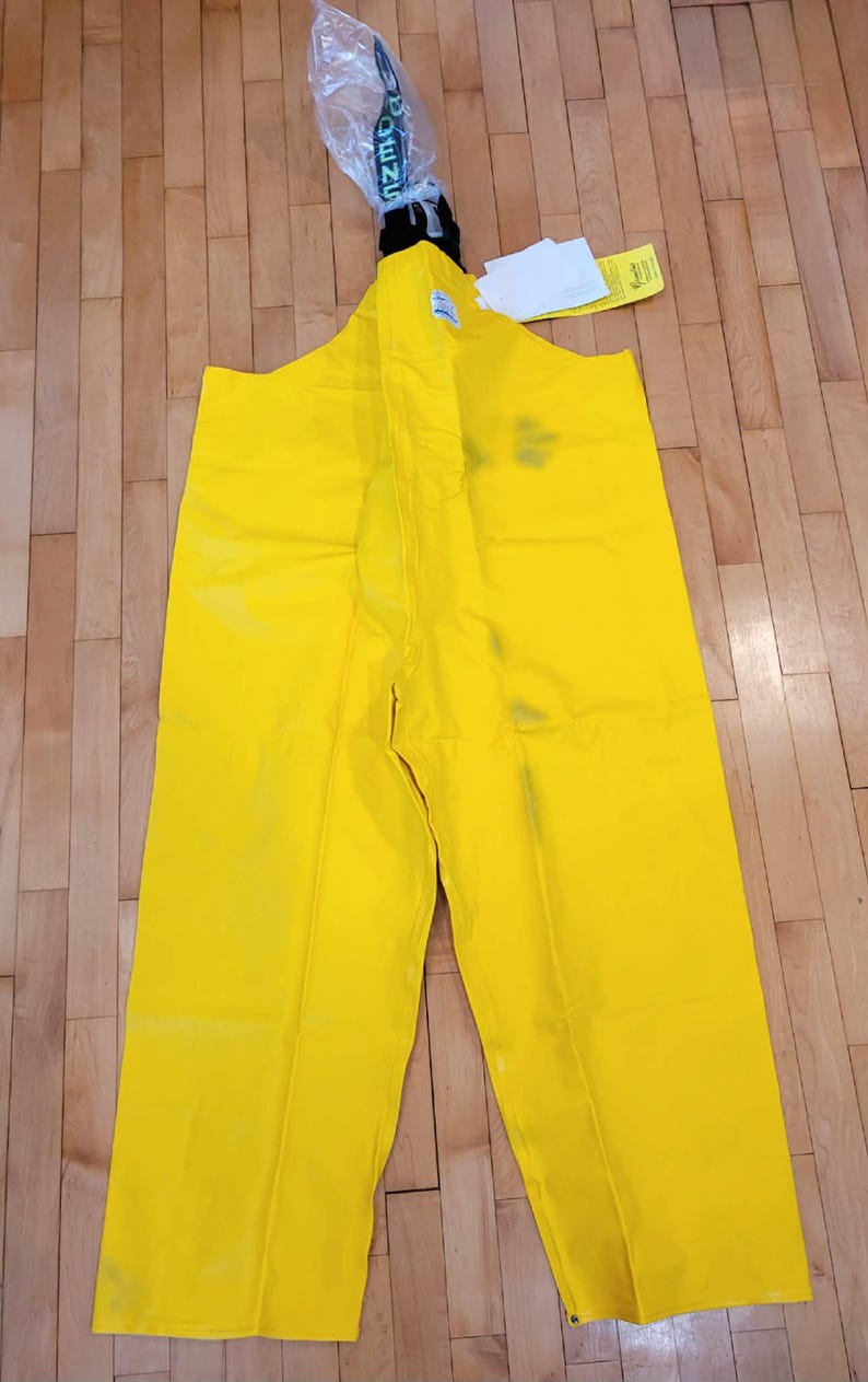 May include: Bright yellow waterproof bib overalls. The overalls have a high bib, suspenders, and straight legs. The garment is made of a shiny, waterproof material. The top of the overalls has a black strap and a clear plastic bag with the word "DENZ" printed on it.