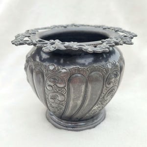 May include: An antique silver-toned vase with a decorative floral rim. The vase has a rounded body with vertical fluting and ornate, embossed floral patterns. The base is circular and slightly raised, adding to its vintage aesthetic.