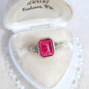 May include: A vintage ring with a large, rectangular, bright pink gemstone set in an ornate, filigree silver band. The ring is displayed in a white jewelry box with the text "HAFF'S JEWELRY, Kaukauna, Wis." on the lid.