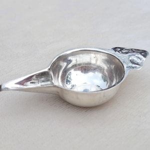 May include: A polished silver-toned metal vessel with a rounded bowl and a long, tapered spout. The handle area features an embossed, intricate decorative pattern. The object has a reflective, metallic surface and a smooth, curved interior basin.