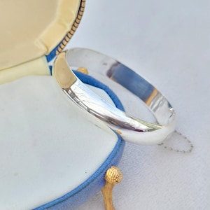 May include: A shiny silver bangle bracelet rests inside a blue and gold jewelry box. The bracelet is smooth and reflective, with a simple, elegant design. The box has a blue velvet exterior and a white satin interior.