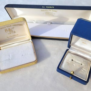 May include: Three vintage jewelry boxes in various colors. A rectangular navy blue box with a white interior and a gold clasp. A gold-colored box with the text "Black Hills Gold Creations" and a cream interior. A long navy box with white interior.