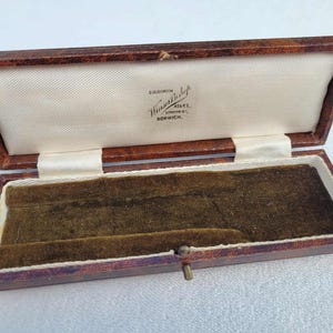 Op de afbeelding: Open antieke bruin lederen sieradendoos met een crèmekleurig satijnen interieur en een donkergroene fluwelen basis. De doos heeft een messing sluiting en de tekst "Goldsmiths & Silversmiths, London & Norwich" is binnenin gedrukt.