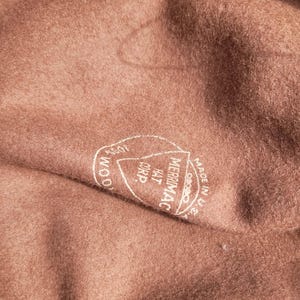 Może przedstawiać: Wnętrze brązowego filcowego kapelusza z białą okrągłą pieczątką. Tekst wewnątrz pieczątki brzmi 100% WOOL, MERRIMAC HAT CORP. oraz MADE IN U.S.A. Tkanina ma miękką, teksturowaną powierzchnię charakterystyczną dla materiału wełnianego.