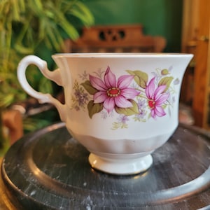 May include: A white teacup with a floral design featuring pink and purple flowers. The cup has a gold rim and a delicate handle.