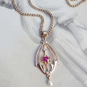 Puede incluir: Un collar con colgante de oro que presenta un marco en forma de lágrima calada, una piedra preciosa rosa redonda y facetada en un engaste con forma de flor, y una pequeña perla blanca colgante en la base, sujeto a una cadena de oro.