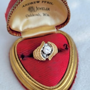 May include: A gold and silver ring with a large, clear diamond in the centre. The ring is in a red velvet heart-shaped box with a gold trim. The box has the inscription "Andrew Pfeil Jeweler Oshkosh, Wis."