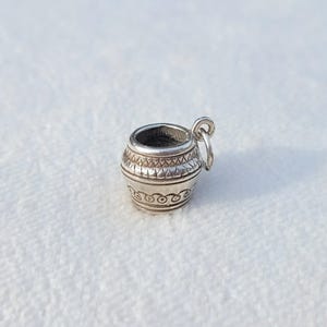 Pode incluir: Um pequeno pingente de prata em forma de vaso ou recipiente arredondado. A superfície metálica apresenta padrões geométricos gravados, incluindo uma faixa de triângulos perto do topo e uma série de motivos circulares na base, com uma pequena argola.