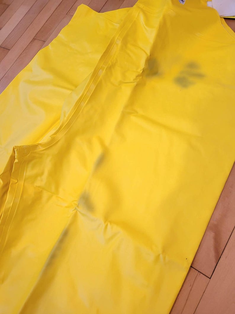May include: Bright yellow waterproof rain pants, likely made of a synthetic material. The pants are folded and lying on a wooden surface. The seams and construction details are visible, suggesting durability and weather protection.