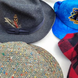 Może przedstawiać: Kolekcja nakryć głowy obejmująca ciemnoszary filcowy kapelusz typu fedora z ciemną wstążką i pi&oacute;rkiem, jaskrawoniebieską czapkę z daszkiem z logo Camel Smooth Character, wielokolorową czapkę z tweedu oraz czerwoną tkaninę w czarną kratę.