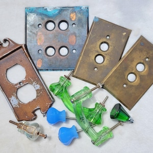 May include: An assortment of vintage electrical hardware, including brown and brass switch plates, and green and blue glass knobs. The switch plates have two or three holes. The knobs are various shapes.