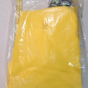 May include: A yellow item is sealed inside a clear plastic bag. The bag has a yellow label on the top and bottom. The item inside appears to be folded. A small portion of a black and green item is visible at the top.