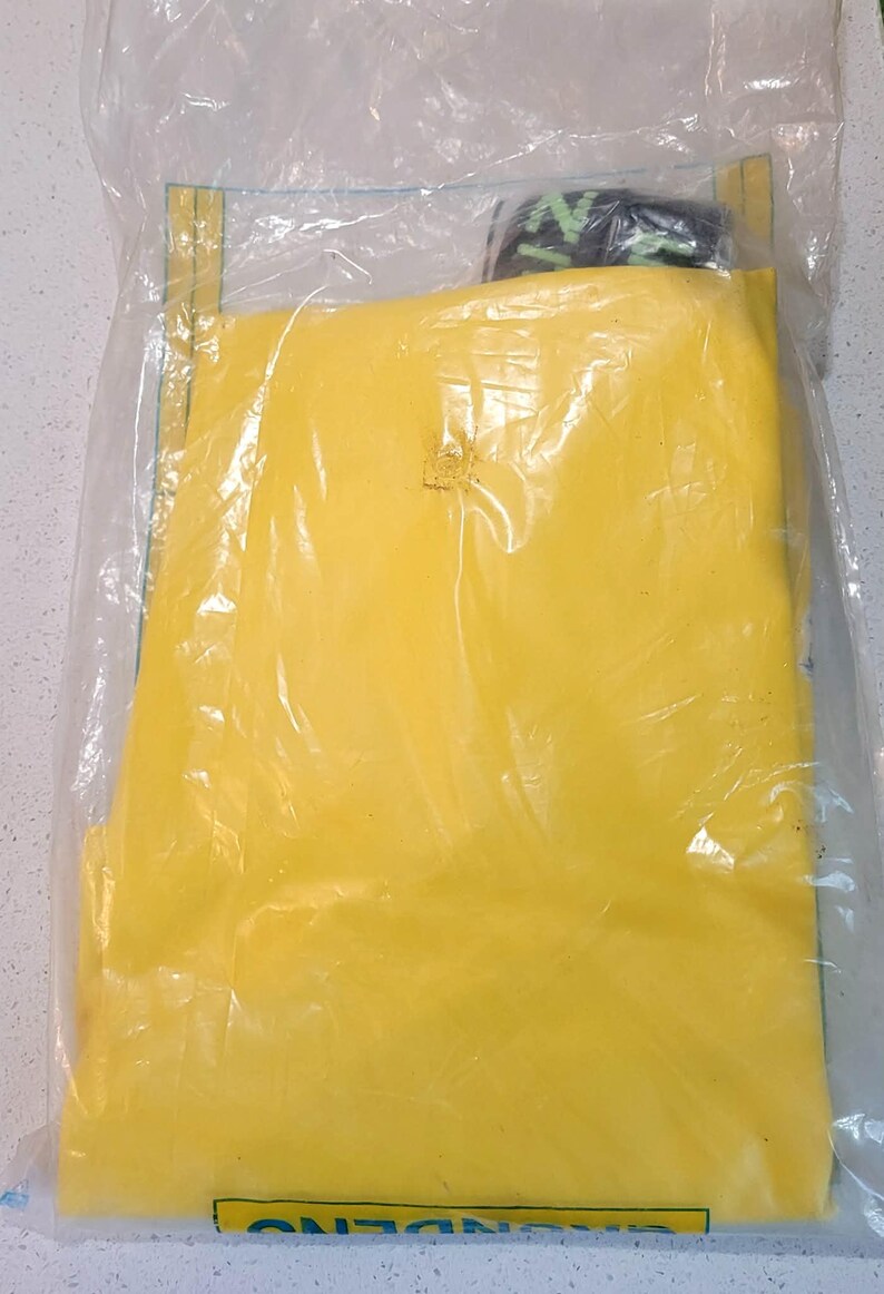 May include: A yellow item is sealed inside a clear plastic bag. The bag has a yellow label on the top and bottom. The item inside appears to be folded. A small portion of a black and green item is visible at the top.