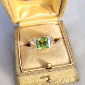 May include: A silver ring featuring a large, square, light green gemstone, presented in an open, vintage jewellery box with a gold interior. The box is tan and has the text "Jewelers Milwaukee, Wis."