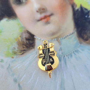 Pode incluir: Um alfinete dourado com um design preto e vermelho. O alfinete apresenta um design de instrumento musical com as letras "IKA" em vermelho. O alfinete é provavelmente um item vintage ou antigo, possivelmente um alfinete de fraternidade ou irmandade.