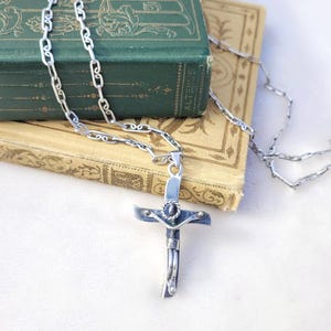 Puede incluir: Un collar con colgante de cruz plateada con un diseño de cruz detallado. La cruz cuelga de una delicada cadena, apoyada sobre un libro verde vintage. El collar se muestra sobre una superficie blanca.