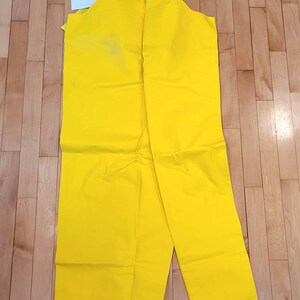 May include: Bright yellow waterproof bib overalls with black straps and a clear plastic bag containing a logo. The overalls are laid out on a wooden floor. A white tag is visible.