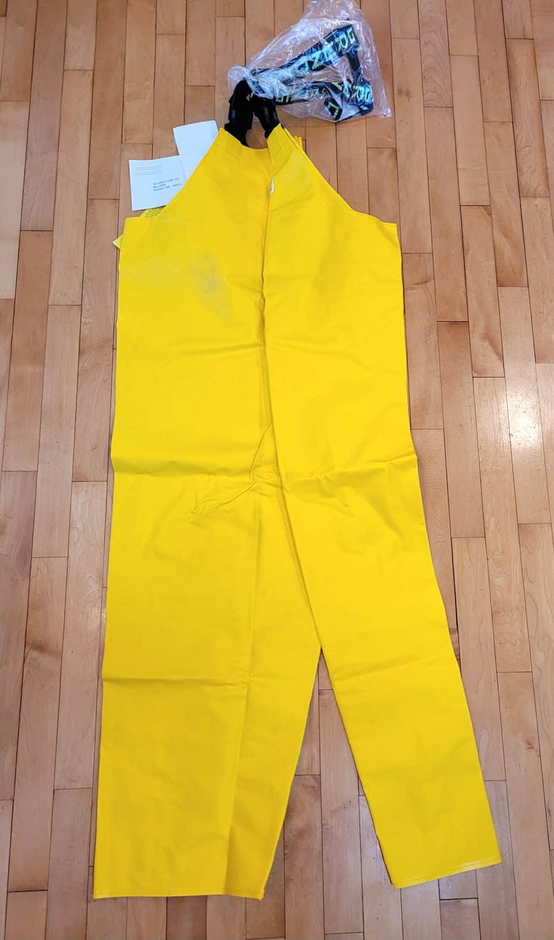May include: Bright yellow waterproof bib overalls with black straps and a clear plastic bag containing a logo. The overalls are laid out on a wooden floor. A white tag is visible.