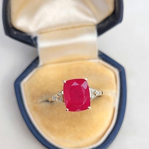Puede incluir: Un anillo de oro con una gran piedra roja de talla cojín sujeta por cuatro garras. La banda incluye pequeñas piedras transparentes engastadas en un diseño metálico decorativo a cada lado. El anillo se presenta dentro de un joyero azul oscuro.