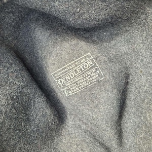 Może przedstawiać: Wnętrze kapelusza z ciemnoszarej filcowanej wełny z białym prostokątnym nadrukiem z tekstem: WARRANTED TO BE A PENDLETON, TRADE MARK REG US PAT OFF, PENDLETON WOOLEN MILLS, PORTLAND OREGON, 100% VIRGIN WOOL. Tkanina ma wyraźnie wyczuwalną teksturę.