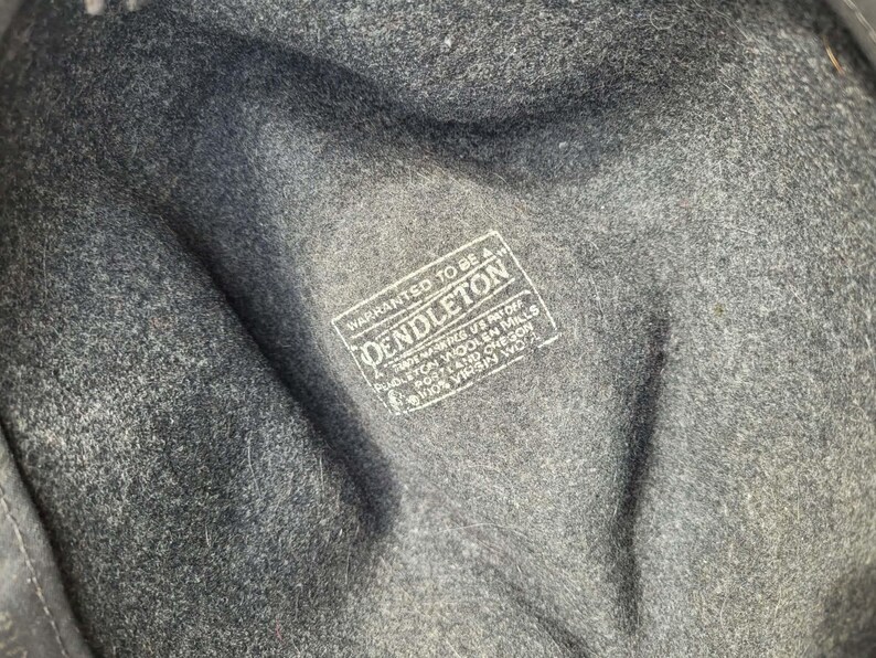 Może przedstawiać: Wnętrze kapelusza z ciemnoszarej filcowanej wełny z białym prostokątnym nadrukiem z tekstem: WARRANTED TO BE A PENDLETON, TRADE MARK REG US PAT OFF, PENDLETON WOOLEN MILLS, PORTLAND OREGON, 100% VIRGIN WOOL. Tkanina ma wyraźnie wyczuwalną teksturę.