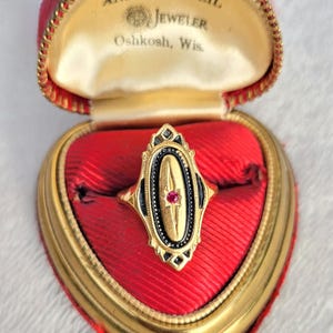 May include: A gold ring with an elongated oval face featuring a black enamel border and a central starburst setting containing a small red gemstone. The ring is presented inside a heart-shaped red velvet jewelry box with gold trim and text.
