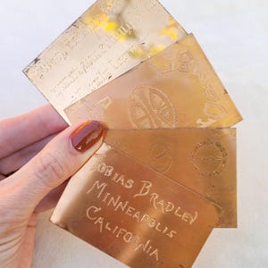 May include: Several rectangular, copper-coloured metal cards are held in a hand. The cards feature etched designs and text. One card reads "Tobias Bradley Minneapolis California". Other cards have circular and geometric designs and text.