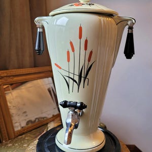 May include: Vintage cream-colored coffee urn with a decorative cattail design in orange and black. Features a spigot, black handles, and a lid with floral accents. The urn sits on a black base, showcasing a retro aesthetic.