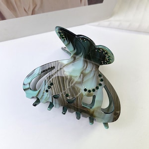 Puede incluir: Una pinza para el pelo en forma de mariposa verde y marr&oacute;n con pedrer&iacute;a. La pinza tiene un patr&oacute;n de espiral y est&aacute; hecha de pl&aacute;stico.