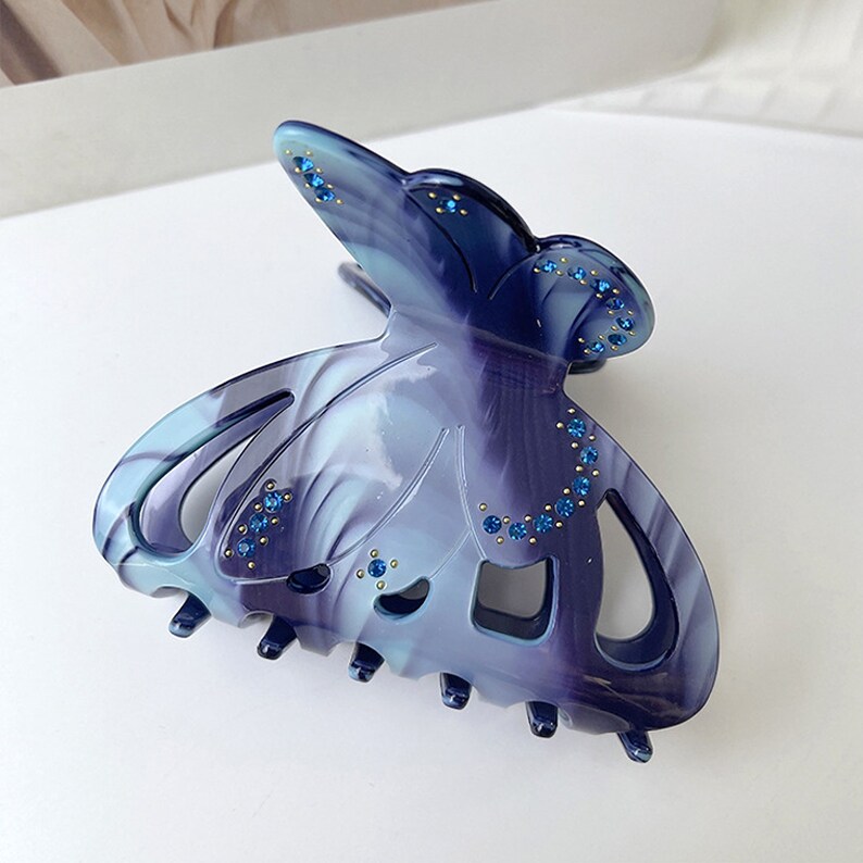 Puede incluir: Una pinza para el pelo en forma de mariposa azul y morada con pedrer&iacute;a. La pinza tiene un patr&oacute;n de espiral y est&aacute; hecha de pl&aacute;stico.