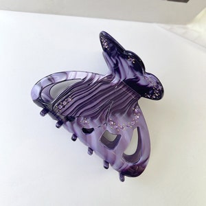 Puede incluir: Pinza para el pelo de color morado en forma de mariposa con un patr&oacute;n de espiral y diamantes de imitaci&oacute;n.