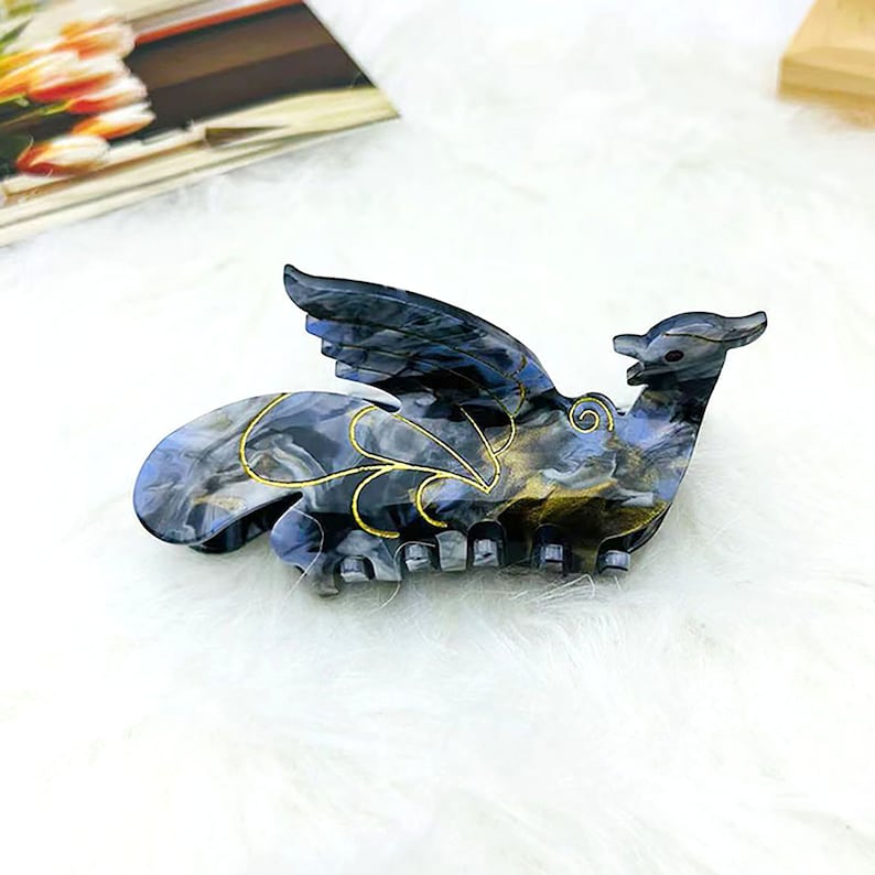 Peut inclure: Une pince &agrave; cheveux noire et dor&eacute;e en forme d'oiseau avec les ailes d&eacute;ploy&eacute;es. La pince a un motif marbr&eacute; et des accents dor&eacute;s.