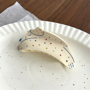 May include: A white plastic hair clip shaped like a cartoon animal with colorful rhinestones and blue eyes.