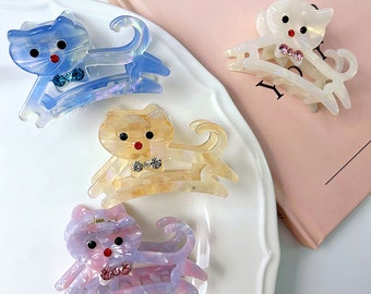 May include: Four colorful cat-shaped hair clips. The clips are made of translucent plastic and have a shiny finish. Each clip has a different color: blue, yellow, pink, and white. Each clip has a small bow and a rhinestone on the front.